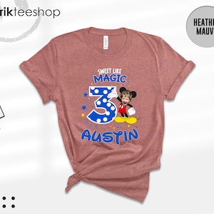 May include: A heather mauve t-shirt featuring a graphic with the text "SWEET LIKE MAGIC" above a large blue polka-dot number 3, a cartoon character with a child's face, and the name "AUSTIN" in blue letters, accented with small yellow stars.
