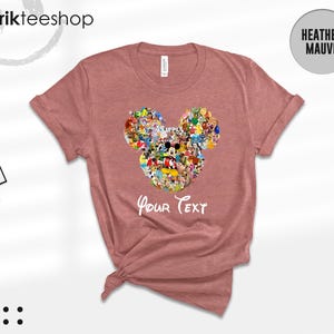 Pu&ograve; includere: T-shirt color malva con un design a forma di testa di Topolino riempita con vari personaggi dei cartoni animati. Le parole "Your Text" sono stampate sotto il disegno. La maglietta ha maniche corte e girocollo.