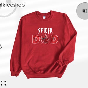 May include: A red crewneck sweatshirt featuring the white text SPIDER DAD on the chest. The letter A is replaced by a small red and blue spider-themed character, and the letters D are filled with a white spiderweb pattern. The brand tag is visible.