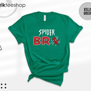 May include: A kelly green short-sleeve t-shirt featuring the white text SPIDER and the red, web-patterned text BRO, with a small red and blue spider-themed character graphic and a white spiderweb design positioned next to the letter O.