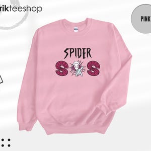 May include: A light pink crewneck sweatshirt featuring a graphic print on the chest. The design includes the text SPIDER above the letters S, O, and S, which are filled with a red spiderweb pattern. A small white spider character is centered.