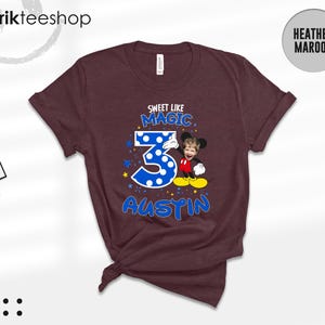 May include: A heather maroon short-sleeve t-shirt featuring a graphic with the text SWEET LIKE MAGIC, the number 3 in blue with white polka dots, and the name AUSTIN. A cartoon character body with a child's face is positioned next to the number 3.