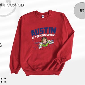 K&ouml;nnte beinhalten: Ein rotes Sweatshirt mit Rundhalsausschnitt und einer Grafik eines Kindes im Weltraum-Ranger-Kost&uuml;m mit Fl&uuml;geln. Der Text AUSTIN IS TURNING SEVEN! ist in blauen Buchstaben gedruckt. Das Design enth&auml;lt kleine blaue Sterne auf der Vorderseite.