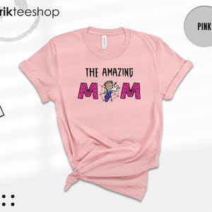 May include: A light pink short-sleeve t-shirt featuring the text THE AMAZING MOM in black letters. The letter O is replaced by a spiderweb graphic with a cartoon character in a blue suit and white gloves positioned in the center of the web.