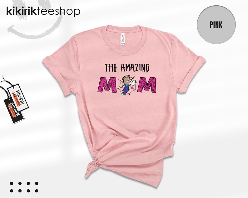 May include: A light pink short-sleeve t-shirt featuring the text THE AMAZING MOM in black letters. The letter O is replaced by a spiderweb graphic with a cartoon character in a blue suit and white gloves positioned in the center of the web.