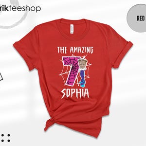 May include: A red short-sleeved t-shirt featuring a large pink number seven with a black spiderweb pattern. The text THE AMAZING is printed above the number, and SOPHIA is printed below. A small photo of a child's face is attached to the number.