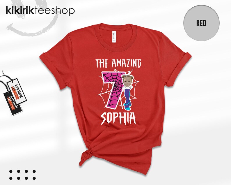May include: A red short-sleeved t-shirt featuring a large pink number seven with a black spiderweb pattern. The text THE AMAZING is printed above the number, and SOPHIA is printed below. A small photo of a child's face is attached to the number.