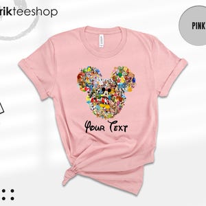 Pu&ograve; includere: T-shirt rosa chiaro con un design a forma di testa di Topolino composto da vari personaggi dei cartoni animati. Le parole "Your Text" sono stampate sotto in nero. La maglietta ha le maniche arrotolate e un'etichetta attaccata.