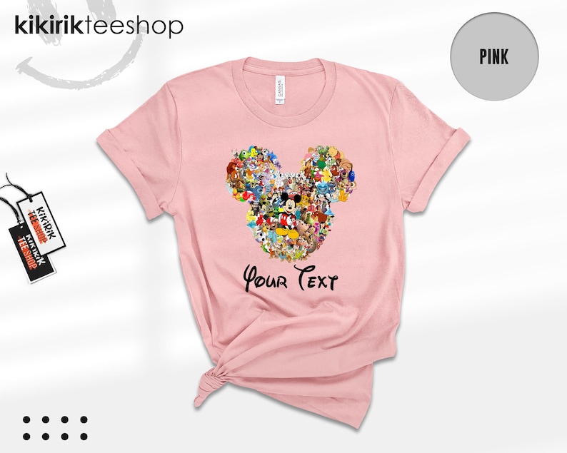 Pu&ograve; includere: T-shirt rosa chiaro con un design a forma di testa di Topolino composto da vari personaggi dei cartoni animati. Le parole "Your Text" sono stampate sotto in nero. La maglietta ha le maniche arrotolate e un'etichetta attaccata.