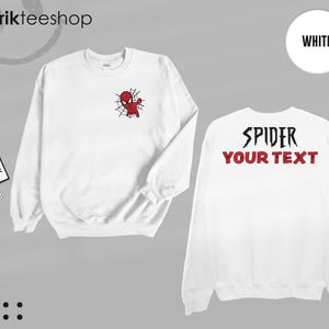 May include: A white long-sleeve sweatshirt featuring a small red and black spider character graphic on the left chest. The back of the sweatshirt displays the black text SPIDER and the red text YOUR TEXT. The garment includes two branded tags.