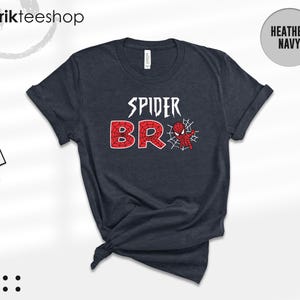 May include: A heather navy short-sleeve t-shirt featuring the white text SPIDER above the red text BRO, with a small red and blue spider-themed character and a white spiderweb graphic integrated into the letter O on the front of the garment.