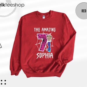 May include: A red crewneck sweatshirt featuring a graphic print with the text THE AMAZING SOPHIA. The design includes a large pink number seven with a black spiderweb pattern, and a small photographic cutout of a child's face.