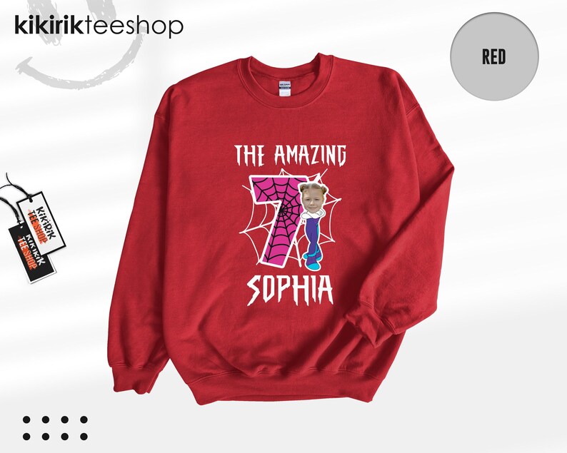 May include: A red crewneck sweatshirt featuring a graphic print with the text THE AMAZING SOPHIA. The design includes a large pink number seven with a black spiderweb pattern, and a small photographic cutout of a child's face.