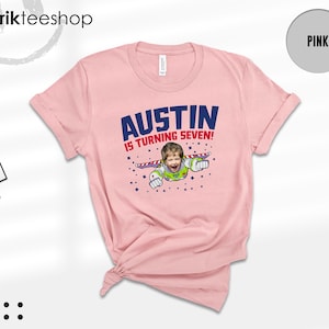 K&ouml;nnte beinhalten: Ein hellrosa T-Shirt mit kurzen &Auml;rmeln und einer Grafik, die das Gesicht eines Kindes auf dem K&ouml;rper einer Weltraum-Ranger-Figur zeigt. Der Text AUSTIN IS TURNING SEVEN! steht in blauen und roten Buchstaben, umgeben von kleinen blauen Sternen.