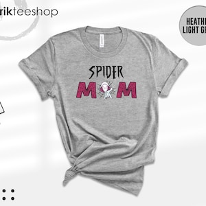 May include: A heather light grey short-sleeve t-shirt featuring the black text SPIDER above the word MOM in pink with a spiderweb pattern. A small white and purple character is positioned between the letters of the word MOM. The shirt has a crew neckline.
