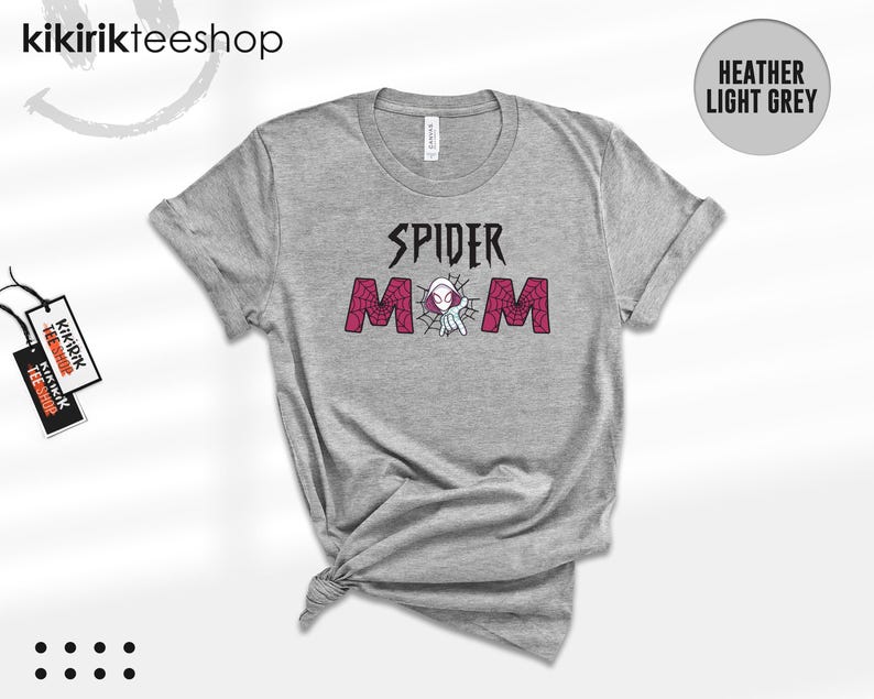 May include: A heather light grey short-sleeve t-shirt featuring the black text SPIDER above the word MOM in pink with a spiderweb pattern. A small white and purple character is positioned between the letters of the word MOM. The shirt has a crew neckline.