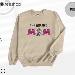 May include: A sand-colored crewneck sweatshirt featuring the text THE AMAZING MOM in black, with the letter O replaced by a spiderweb graphic containing a small cartoon character in a blue outfit and white gloves, and a tag reading kikirikteeshop.