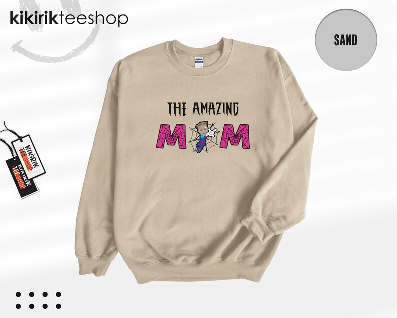 May include: A sand-colored crewneck sweatshirt featuring the text THE AMAZING MOM in black, with the letter O replaced by a spiderweb graphic containing a small cartoon character in a blue outfit and white gloves, and a tag reading kikirikteeshop.