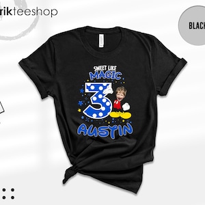May include: A black short-sleeve t-shirt featuring a graphic print with the text "SWEET LIKE MAGIC" and "AUSTIN". The design includes a large blue polka-dot number 3, a cartoon character body with yellow shoes, and a child's face photo.