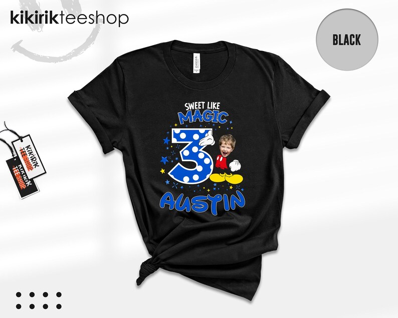 May include: A black short-sleeve t-shirt featuring a graphic print with the text "SWEET LIKE MAGIC" and "AUSTIN". The design includes a large blue polka-dot number 3, a cartoon character body with yellow shoes, and a child's face photo.
