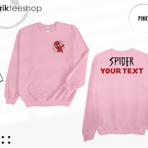 May include: A light pink crewneck sweatshirt featuring a small graphic of a red spider-themed character on a web on the left chest. The back displays the black text SPIDER and the red text YOUR TEXT. A tag with the text KIKIRIKTEESHOP is attached.