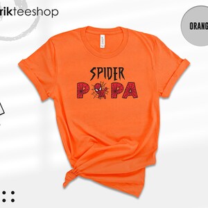 Puede incluir: Camiseta naranja brillante con las palabras "SPIDER PA" en negro y rojo, con una telara&ntilde;a y un gr&aacute;fico de ara&ntilde;a. La camiseta tiene las mangas enrolladas y una etiqueta que dice "kikirikteeshop".