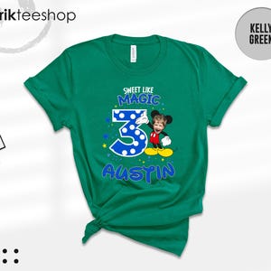 May include: A kelly green short-sleeve t-shirt featuring a graphic with the text "SWEET LIKE MAGIC" above a large blue polka-dot number "3" and the name "AUSTIN" below. A cartoon character with a child's face is positioned next to the number.