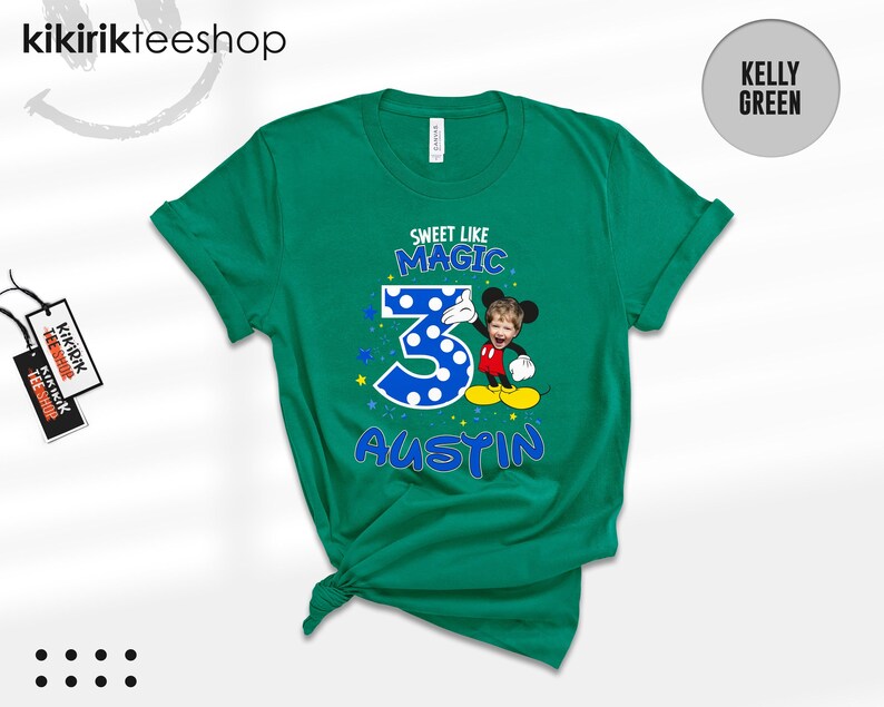 May include: A kelly green short-sleeve t-shirt featuring a graphic with the text "SWEET LIKE MAGIC" above a large blue polka-dot number "3" and the name "AUSTIN" below. A cartoon character with a child's face is positioned next to the number.