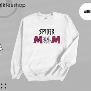 May include: A white long-sleeve crewneck sweatshirt featuring the text SPIDER MOM in black and red. The letter O is replaced by a graphic of a white and purple spider-themed character inside a spider web. The sweatshirt has a ribbed collar and cuffs.