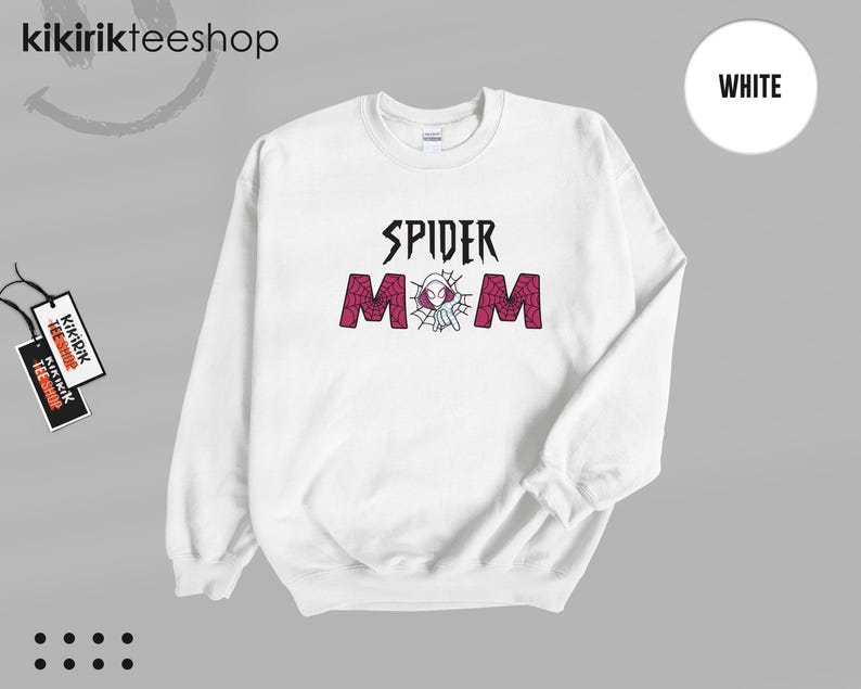 May include: A white long-sleeve crewneck sweatshirt featuring the text SPIDER MOM in black and red. The letter O is replaced by a graphic of a white and purple spider-themed character inside a spider web. The sweatshirt has a ribbed collar and cuffs.