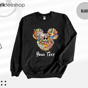 Pu&ograve; includere: Felpa girocollo nera con un colorato design a forma di testa di Topolino composto da vari personaggi dei cartoni animati. Le parole "Your Text" sono scritte in bianco sotto. La felpa ha un'etichetta con scritto "Kikirik Tees Shop".