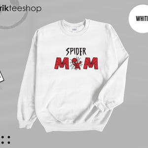 Puede incluir: Sudadera blanca con el texto "SPIDER MOM" en negro y rojo. El dise&ntilde;o presenta una telara&ntilde;a y una ara&ntilde;a de dibujos animados. La sudadera tiene cuello redondo y mangas largas. La marca "kikirikteeshop" es visible en la esquina superior izquierda.