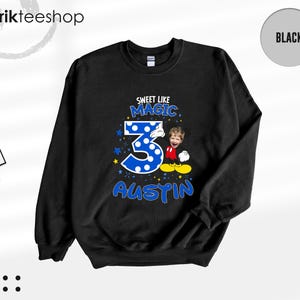 May include: A black long-sleeve sweatshirt featuring a graphic print with the text "SWEET LIKE MAGIC" above a large blue polka-dot number three, the name "AUSTIN" below, and a cartoon character body with a child's face and yellow shoes.