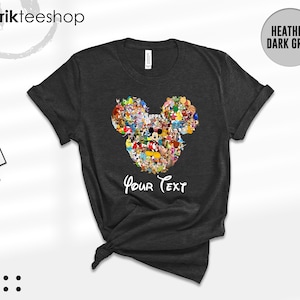 Pu&ograve; includere: T-shirt grigio scuro con un design a forma di testa di Topolino riempito con vari personaggi dei cartoni animati. Le parole "Your Text" sono scritte in un carattere script sottostante. La maglietta ha le maniche arrotolate.