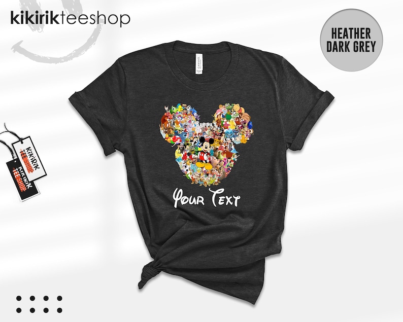 Pu&ograve; includere: T-shirt grigio scuro con un design a forma di testa di Topolino riempito con vari personaggi dei cartoni animati. Le parole "Your Text" sono scritte in un carattere script sottostante. La maglietta ha le maniche arrotolate.