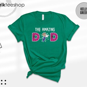 May include: A kelly green short-sleeve t-shirt featuring the text THE AMAZING DAD in white. The letter A is replaced by a small cartoon figure in a blue and purple suit, and the letters D are filled with a pink spiderweb pattern.