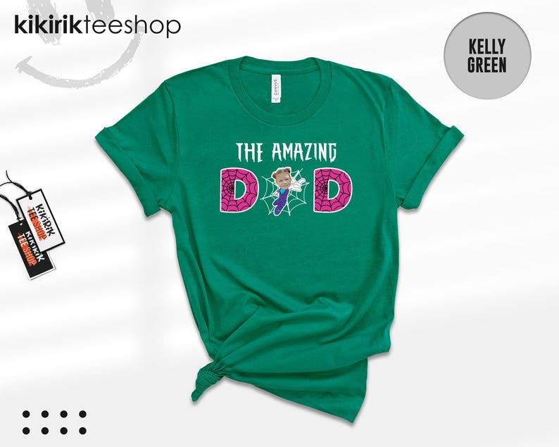 May include: A kelly green short-sleeve t-shirt featuring the text THE AMAZING DAD in white. The letter A is replaced by a small cartoon figure in a blue and purple suit, and the letters D are filled with a pink spiderweb pattern.