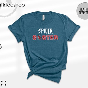 Puede incluir: Camiseta de color verde azulado oscuro con las palabras "SPIDER SISTER" en letras blancas y rojas. Un peque&ntilde;o gr&aacute;fico de una ara&ntilde;a est&aacute; entre las palabras. La camiseta est&aacute; anudada en la parte inferior.