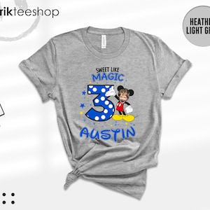 May include: A heather light grey short-sleeve t-shirt featuring a graphic print with the text "SWEET LIKE MAGIC" above a large blue polka-dot number three, the name "AUSTIN" below, and a cartoon character with a child's face photo.