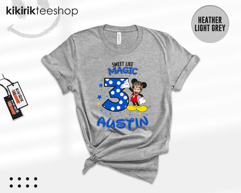 May include: A heather light grey short-sleeve t-shirt featuring a graphic print with the text "SWEET LIKE MAGIC" above a large blue polka-dot number three, the name "AUSTIN" below, and a cartoon character with a child's face photo.