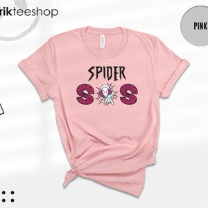 May include: A light pink short-sleeve t-shirt featuring the text SPIDER SOS in black and dark red. The letter O is replaced by a graphic of a white and purple spider-themed character in a web. The shirt has a crew neckline and a knotted hem.