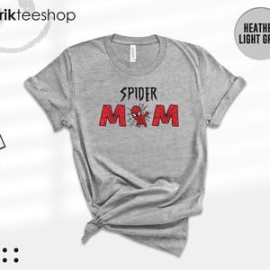 Puede incluir: Camiseta gris jaspeado claro con las palabras "SPIDER MOM" en negro y rojo. El dise&ntilde;o incluye un gr&aacute;fico de ara&ntilde;a. La camiseta est&aacute; anudada en la parte inferior. La marca "kikirikteeshop" es visible en la parte superior izquierda.