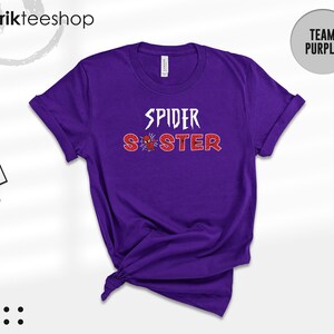 Puede incluir: Una camiseta morada con las palabras "SPIDER SISTER" en blanco y rojo, con un gr&aacute;fico de ara&ntilde;a. La camiseta tiene las mangas enrolladas y un nudo en la parte inferior. Una etiqueta de la tienda est&aacute; adjunta. El texto "TEAM PURPLE" est&aacute; en un c&iacute;rculo.