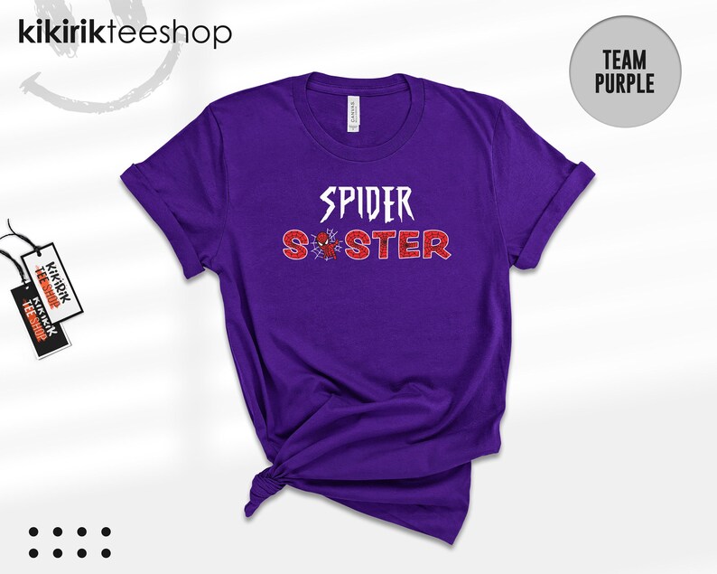Puede incluir: Una camiseta morada con las palabras "SPIDER SISTER" en blanco y rojo, con un gr&aacute;fico de ara&ntilde;a. La camiseta tiene las mangas enrolladas y un nudo en la parte inferior. Una etiqueta de la tienda est&aacute; adjunta. El texto "TEAM PURPLE" est&aacute; en un c&iacute;rculo.