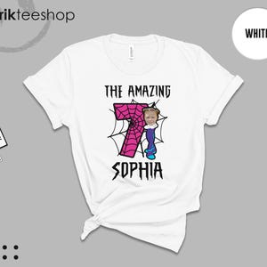 May include: A white short-sleeve t-shirt featuring a graphic of a large pink number seven with a spiderweb pattern. The design includes the text THE AMAZING SOPHIA and a small cartoon character with a photo-realistic face attached to the number.