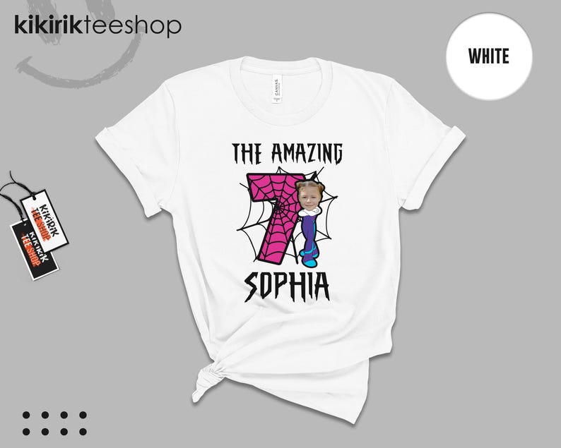 May include: A white short-sleeve t-shirt featuring a graphic of a large pink number seven with a spiderweb pattern. The design includes the text THE AMAZING SOPHIA and a small cartoon character with a photo-realistic face attached to the number.