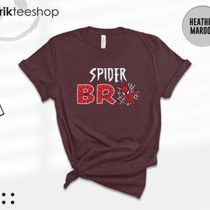 May include: A heather maroon short-sleeve t-shirt featuring the white text SPIDER above the red web-patterned text BRO, with a small red and blue spider-themed character graphic positioned next to the letter O and a white spider web design.