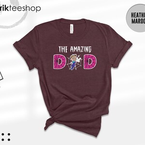 May include: A heather maroon short-sleeve t-shirt featuring the text THE AMAZING DAD in white letters. The letter A is replaced by a graphic of a person in a blue and purple suit, and the two Ds are filled with a pink spider web pattern.