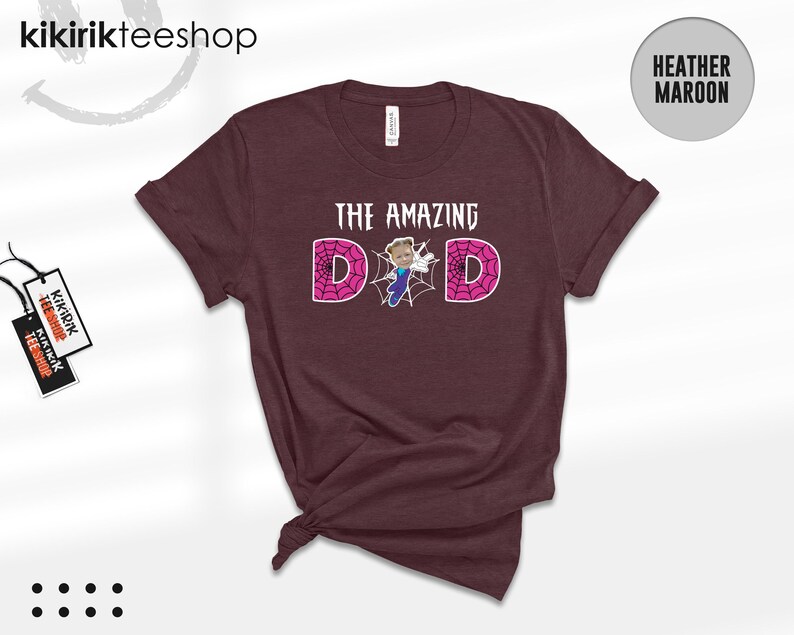 May include: A heather maroon short-sleeve t-shirt featuring the text THE AMAZING DAD in white letters. The letter A is replaced by a graphic of a person in a blue and purple suit, and the two Ds are filled with a pink spider web pattern.