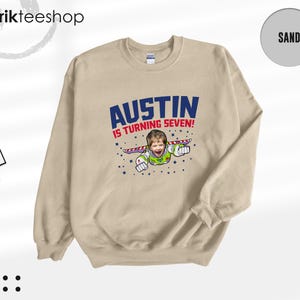 K&ouml;nnte beinhalten: Ein sandfarbenes Sweatshirt mit Rundhalsausschnitt und einem Grafikdruck, der das Gesicht eines Kindes auf dem K&ouml;rper eines Weltraum-Rangers zeigt. Der Text AUSTIN IS TURNING SEVEN! ist in blauen und roten Buchstaben mit Sternen abgebildet.